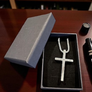 May include: A silver cross pendant with the inscription "HEBREWS 12:29" on a chain, presented in a grey gift box. A black and gold pen and a small black object are also visible on a dark wooden surface.