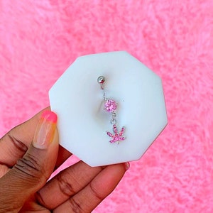 Pode incluir: Um piercing corporal de prata com um cristal rosa e um amuleto de folha de maconha de cristal rosa.