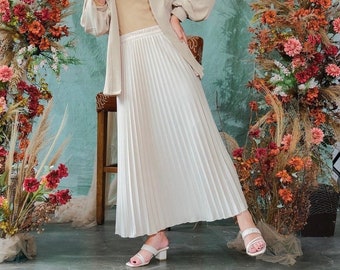 May include: A woman wearing a white headscarf, a beige cardigan, a beige top, and a white pleated skirt. She is standing in front of a wall with a floral arrangement on either side.