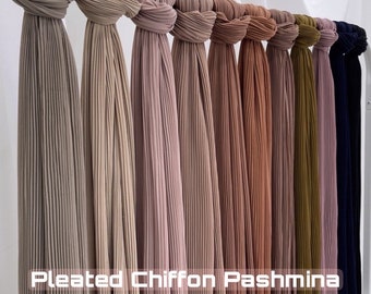 May include: A collection of pleated chiffon pashminas in various colors, including beige, brown, pink, peach, and green. The pashminas are arranged in a row, hanging from a rod. The text "Pleated Chiffon Pashmina" and "by AdhaLabels" is visible at the bottom of the image.