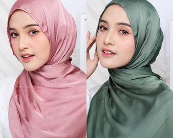 May include: Two women wearing satin hijabs in a studio setting. One hijab is a dusty rose color, and the other is a muted green. The hijabs are draped elegantly, with soft folds and a subtle sheen. The brand name "Adha Labels" is printed on the bottom of the image.