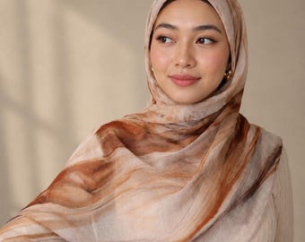May include: A lightweight hijab featuring a swirling abstract pattern in shades of warm brown, cream, and tan. The fabric has a soft, semi-sheer texture with fluid, marbled lines that drape across the shoulders and wrap around the head.