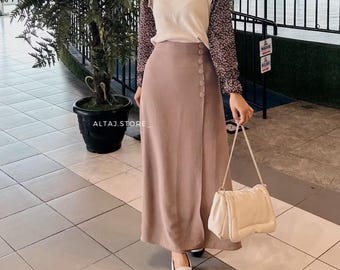 May include: A woman wearing a long, brown skirt with a white top and a floral print blouse. She is carrying a white handbag with a chain strap. The skirt has a button closure and a slit on the side.