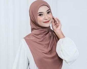May include: A mocha-colored hijab, draped elegantly, covers the head and shoulders. The fabric has a smooth, matte finish. The hijab is paired with a white blouse. The text "Adha Labels" is visible on the hijab.
