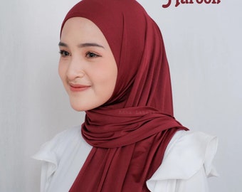 May include: A maroon hijab, draped over the head and shoulders. The fabric is a solid, deep red color. The word "Maroon" is displayed in a script font. The hijab is worn over a white blouse.