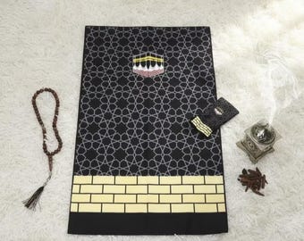 May include: Black prayer rug with a geometric pattern and a gold brick design at the bottom. The rug features an image of the Kaaba. A small black pouch, prayer beads, and a decorative incense burner are also present.