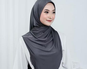 May include: A dark gray hijab, draped elegantly. The fabric appears smooth and soft, with subtle folds. The text "Adha Labels" is visible on the garment. The hijab is worn over a white garment.