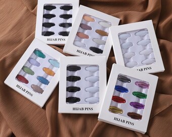 May include: Six white rectangular display cards, each containing ten oval-shaped hijab pins in various colors. The cards are labeled HIJAB PINS in black text. The pins are arranged in two vertical columns of five on each card, featuring solid, metallic, and pastel finishes.