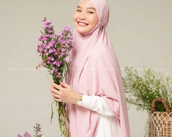 May include: A woman wearing a light pink hijab and white shirt, holding a bouquet of purple flowers. The image has the text "Adha Labels" repeated across the bottom. The woman is smiling.