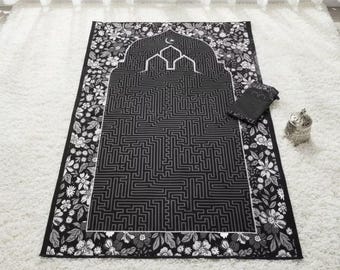 May include: Black and white prayer rug with a floral border. The center features a maze-like design with an arched top and a crescent moon. A matching prayer mat and a small silver container are also visible.