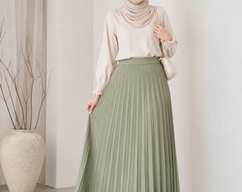 May include: A long, sage green pleated skirt with a high waistband, paired with a long-sleeved beige blouse featuring gathered cuffs and a matching beige hijab, worn with pointed-toe beige shoes and a small beige shoulder bag.