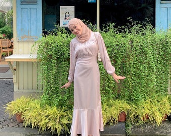 May include: A light pink satin dress with long sleeves and a mermaid hem. The dress has a fitted waist and a round neckline. The person in the image is wearing a matching headscarf and silver sandals. A white purse with a pearl strap is also visible.