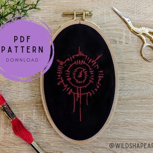 Peut inclure: Un motif de broderie rouge sur un tissu noir dans un cerceau de broderie en bois. Le motif est un cercle avec un design en spirale et une ligne verticale au centre. Le cerceau est sur une surface en bois avec une bobine de fil à broder rouge et une paire de ciseaux dorés.