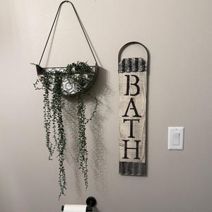 May include: A black metal hanging planter with a honeycomb pattern and trailing green plants. Next to it is a vertical wooden sign with the word "BATH" in black letters, and a metal handle.