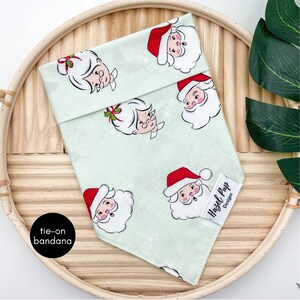May include: A green tie-on bandana with a white background and a repeating pattern of Santa Claus and Mrs. Claus faces. The bandana has a label that says "Hazel Pflug Designs".
