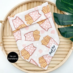 May include: A pink and white tie-on bandana for a pet with a pattern of french fries in pink boxes with pink hearts. The bandana has a white tag that says "Hazel Pup Designs".