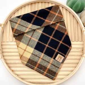 May include: A black, brown, and tan plaid bandana with a brown leather tag that says "Hazel & The Dogs".