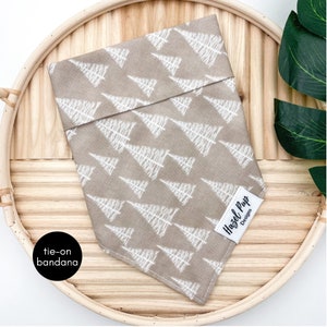 May include: A tan tie-on bandana with a white tree pattern. The bandana has a white label that says "Hazel Pup Designs".