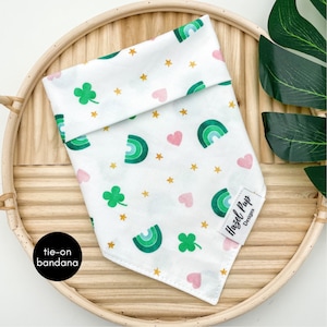May include: White tie-on bandana with a green shamrock, rainbow, and pink heart pattern. The bandana has gold stars and the text "Hazel Pup Designs" in black.