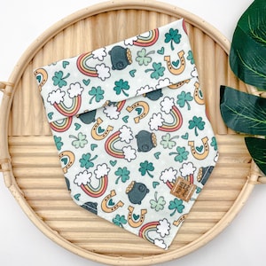 Op de afbeelding: Een groene, witte en oranje hondenbandana met een patroon van regenbogen, klavertjesvier, gouden potten en hoefijzers. De bandana heeft een bruin label met de tekst "Hazel & Dewey Designs".