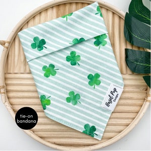 May include: A green and white tie-on bandana with a shamrock pattern. The bandana is on a woven basket. The text "Hazel Pup Designs" is on the bandana.