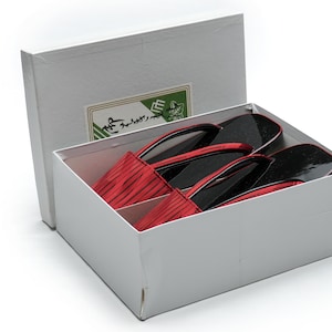 May include: A pair of red and black Japanese sandals, called geta, in a white box with a green label. The sandals have a wooden base and a red fabric upper.