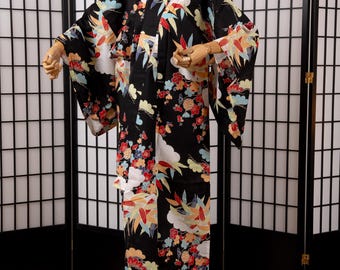 May include: A black kimono featuring a vibrant, multi-colored pattern of stylized flowers, leaves, and cloud-like shapes in shades of red, blue, yellow, and white. The garment has long, wide sleeves and a full-length silhouette with a wrap-front design.