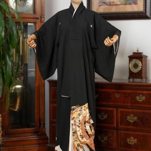 May include: A black kimono with a white collar and cuffs, displayed on a mannequin. The lower portion features a decorative panel with floral and geometric patterns in shades of gold, red, and cream. The garment is a traditional Japanese robe.