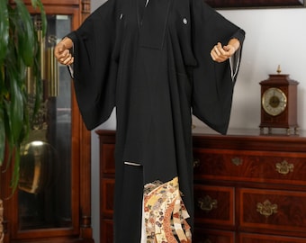 May include: A black kimono with a white collar and cuffs, displayed on a mannequin. The lower portion features a decorative panel with floral and geometric patterns in shades of gold, red, and cream. The garment is a traditional Japanese robe.
