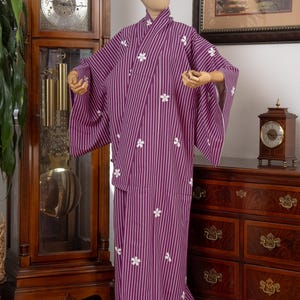 May include: A purple kimono with vertical white stripes and white floral accents. The kimono is displayed on a mannequin. The garment has wide sleeves and a wrap-around design. The background includes a grandfather clock and a wooden dresser.
