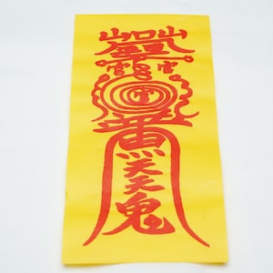 May include: A yellow paper with red Chinese characters printed on it. The characters are in a vertical column and are surrounded by a red circle. The paper is folded in half.