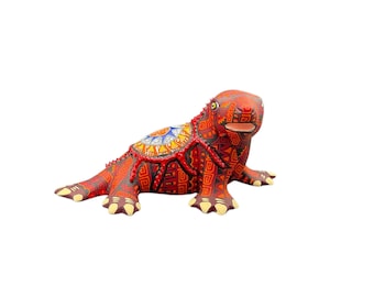 May include: A handcrafted, colorful lizard figurine. The sculpture features intricate patterns in red, orange, and brown, with accents of yellow, blue, and white. The lizard is posed on all fours, showcasing its detailed design.