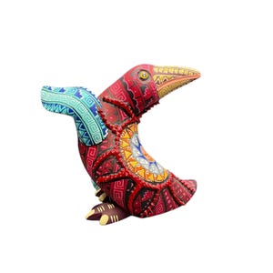 Puede incluir: Una escultura de pájaro pintada con colores vibrantes, con cuerpo rojo, ala azul y pico amarillo. La pieza de arte popular presenta intrincados patrones geométricos en varios colores, incluyendo rojo, azul, amarillo y naranja. El pájaro está sobre un fondo blanco.