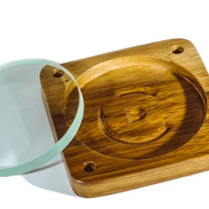 May include: A wooden coaster with a circular indentation and a clear, round glass lens. The coaster is square with rounded corners and four small holes. The wood has a warm, natural tone.