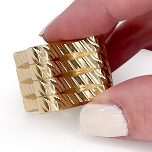 May include: A gold-colored ring with a textured, geometric design. The ring is held between two fingers, showcasing its intricate pattern. The ring's surface reflects light, highlighting its detailed craftsmanship. The ring appears to be a fashion accessory.