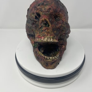 May include: A grotesque, decaying skull with exposed teeth and an open mouth. The skull is painted in shades of brown, red, and green, with a textured, weathered appearance. The skull is a Halloween decoration.