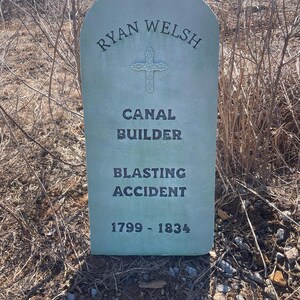 May include: A weathered green tombstone with the inscription "Ryan Welsh, Canal Builder, Blasting Accident, 1799 - 1834". A cross is carved above the inscription.