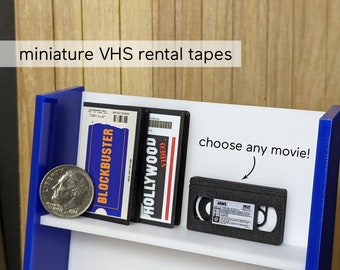 May include: A miniature shelf with three VHS tapes, two with labels that say "Blockbuster" and "Hollywood Video", and one with a black label that says "Jaws". The shelf is blue and white and has the text "choose any movie!" written on it.
