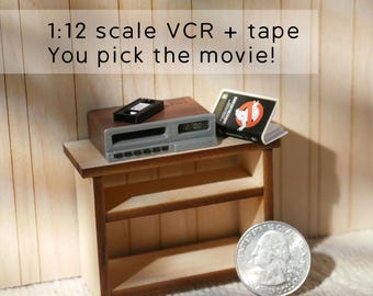 May include: Miniature VCR and tape set with the text "1:12 scale VCR + tape You pick the movie!". The VCR is brown and gray, with a black tape and a Ghostbusters movie tape. The set is on a small wooden shelf, with a quarter for scale.