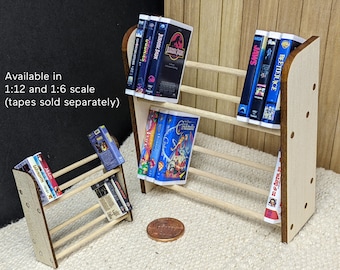 May include: Two wooden miniature bookshelves with shelves for holding VHS tapes. The shelves are filled with various VHS tapes with titles like "Jaws", "Beetlejuice", "Jurassic Park", and "The Goonies". The shelves are available in 1:12 and 1:6 scale. Tapes sold separately.