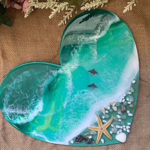 Puede incluir: Una bandeja de resina en forma de corazón con un diseño de ola de océano verde y blanco. La bandeja presenta una estrella de mar, conchas marinas y pequeñas piedras blancas.