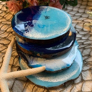 Puede incluir: Conjunto de cuatro posavasos de resina azul y blanca con base de madera. Los posavasos presentan un diseño de ola con espuma blanca y una pequeña tortuga marina.