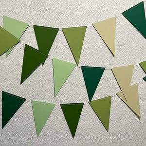 May include: A garland of triangular flags in various shades of green and cream hangs against a white textured wall. The flags are strung together, creating a festive and decorative banner. The color palette is natural and earthy.