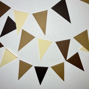 May include: A decorative banner with triangular flags in shades of brown and cream. The flags are strung together on a thin white string, creating a festive garland. The banner is against a white wall.