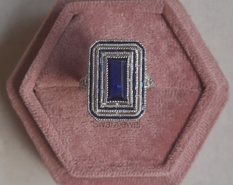 May include: A silver ring with a large, rectangular, dark blue gemstone surrounded by a decorative silver frame. The ring is in a pink velvet ring box.
