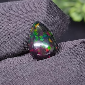 May include: A teardrop-shaped black opal gemstone with vibrant flashes of green, red, and yellow. The polished stone is held against a dark background, showcasing its iridescent play-of-color. This gemstone is a unique piece of jewelry.