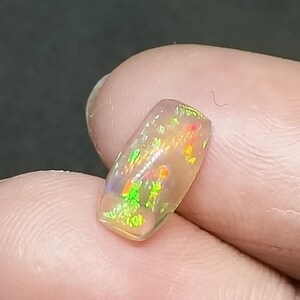 May include: A small, rectangular, iridescent opal gemstone with a light pink base and green, orange, and red flashes of color.