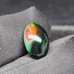 May include: A polished gemstone with a mix of colors, including green, orange, and black. The oval-shaped stone is held against a dark gray fabric, showcasing its vibrant hues and glossy surface. The gemstone's unique color pattern is the focal point.