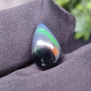 May include: A teardrop-shaped, polished gemstone with a dark base color. The stone reflects a vibrant spectrum of colors, including blue, green, and orange, creating a shimmering effect. The gemstone is held against a dark gray fabric, with greenery in the background.
