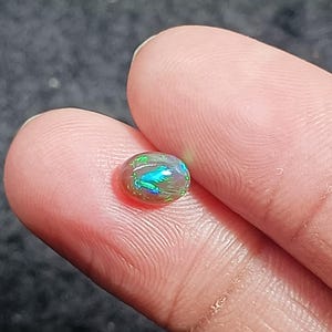 Könnte beinhalten: Ein ovaler Opal-Edelstein in Dunkelgrau mit grün-blauen, schillernden Blitzen.